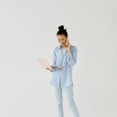 Asian woman in blue attire multitasking with a laptop and phone call.